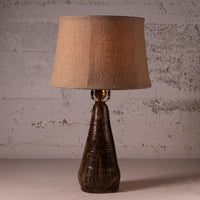 A rustic table lamp with a textured brown ceramic base and a natural beige linen drum shade, set against a distressed concrete wall.