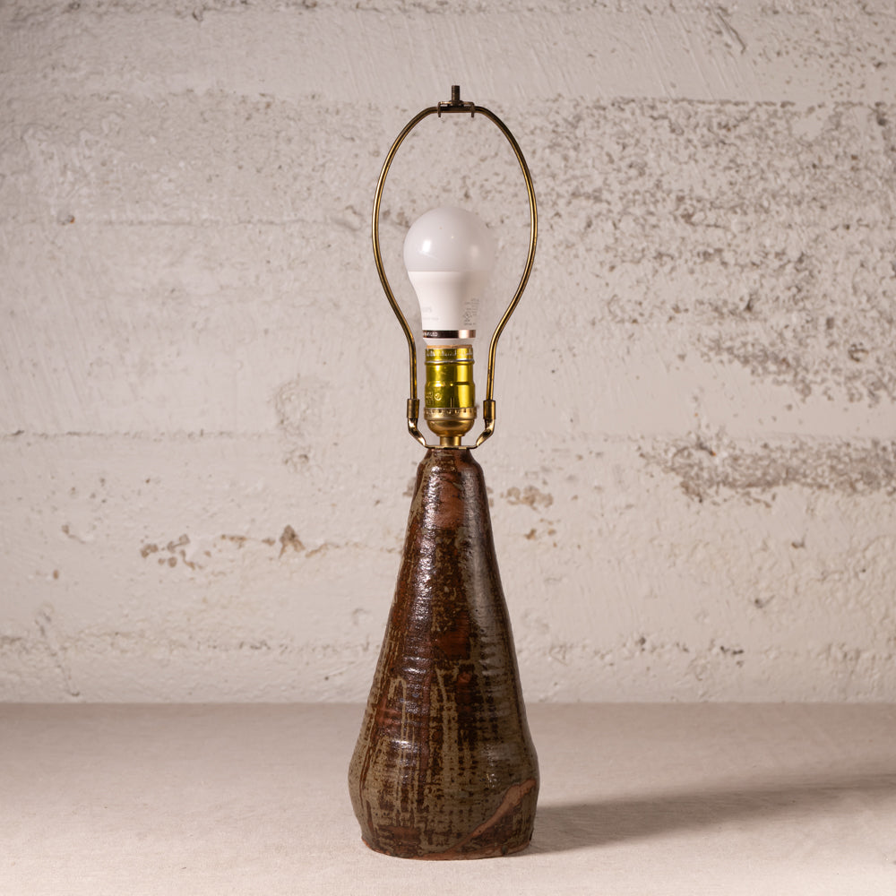 A rustic table lamp with a textured, tapered dark brown ceramic base and a brass-colored metal arched shade holding a white bulb, set against a distressed off-white brick wall.