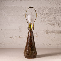 A rustic table lamp with a textured, tapered dark brown ceramic base and a brass-colored metal arched shade holding a white bulb, set against a distressed off-white brick wall.