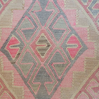 A close-up of a woven textile with a geometric kilim-style pattern in pink, beige, and gray.