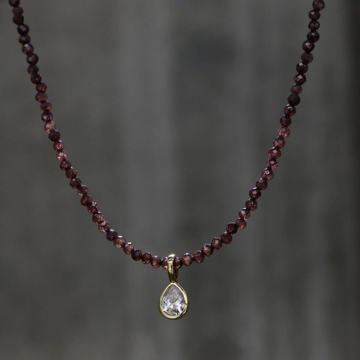 A beaded necklace with small dark red garnet-like beads and a teardrop-shaped clear stone pendant in a gold setting, shown against a blurred gray background.