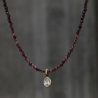 A beaded necklace with small dark red garnet-like beads and a teardrop-shaped clear stone pendant in a gold setting, shown against a blurred gray background.