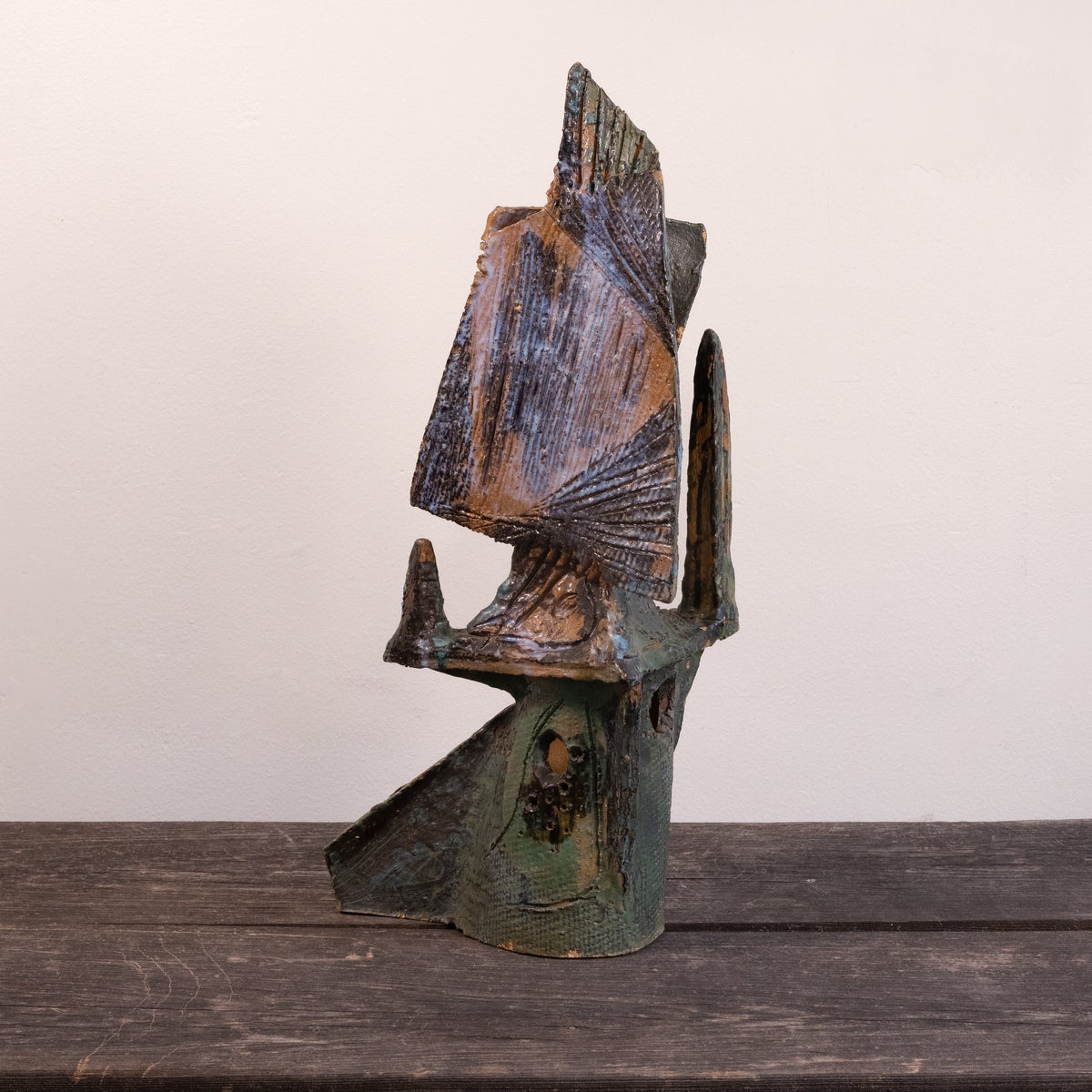 A rustic, handcrafted ceramic sculpture of an abstract figure, featuring weathered brown and green patina textures, displayed on a dark wooden surface against a plain off-white wall.