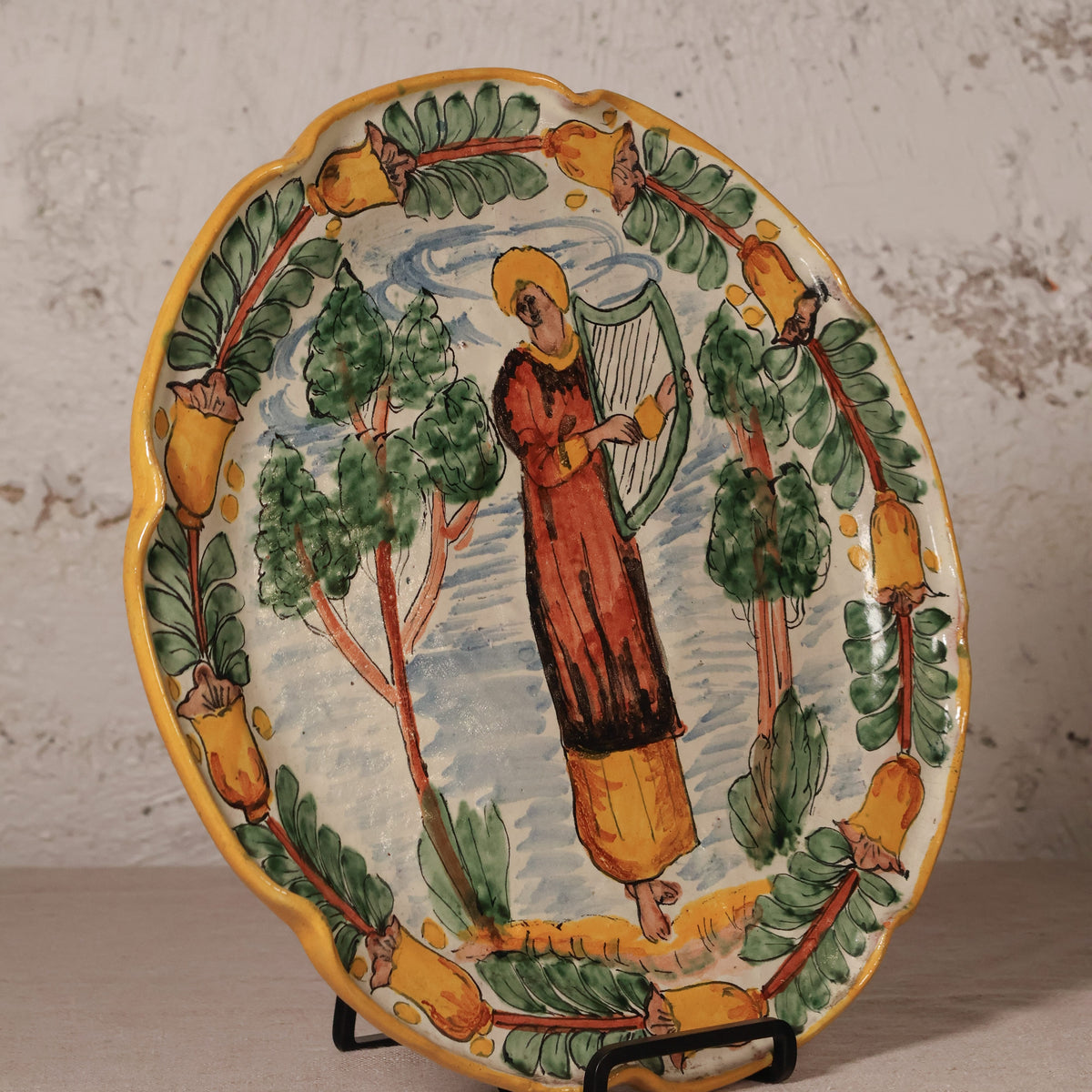Ceramic plate with a decorative design of a person playing a harp surrounded by trees and fruit on a stand.
