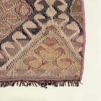 Close-up of a woven rug corner showing a geometric pattern in beige, brown, muted pink, and dark charcoal tones with a fringed edge on a light cream background.