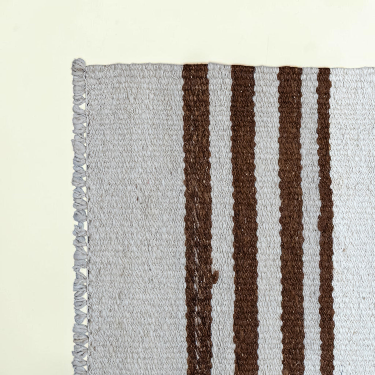 Close-up of a woven textile corner with light gray fringed edges and vertical dark brown stripes on a pale background.