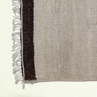 Close-up of a woven rug corner showing a light beige main field with a dark brown vertical stripe and fringed edge on a white background.