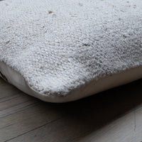 A close-up of a textured off-white sheepskin or wool throw pillow resting on a dark wood floor.