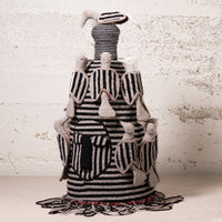 A handmade textile sculpture of a rooster, featuring black and white striped fabric with beige feather details, set against a textured off-white brick wall.