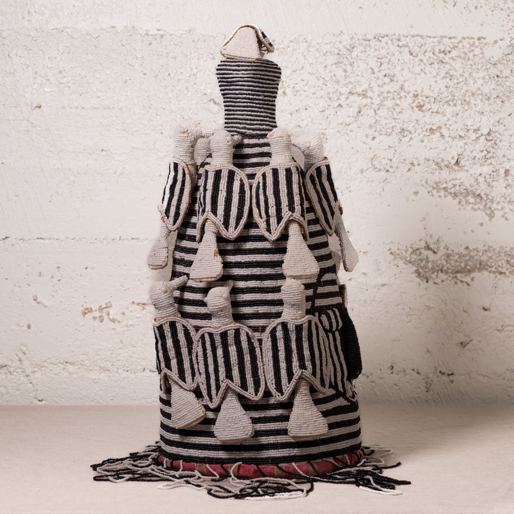 A handwoven Moroccan-style decorative basket or vase with a black and beige striped pattern, featuring textured geometric details, tassels, and fringe, displayed against a rustic white plaster wall.