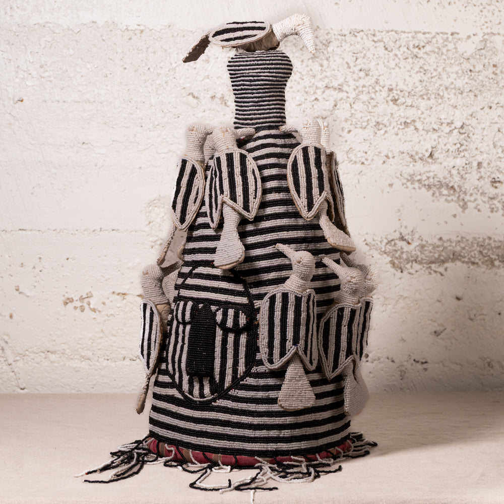 A handmade textile doll or decorative figure with black and white striped fabric, beige felted wool details, and hanging tassels, set against a textured off-white wall.