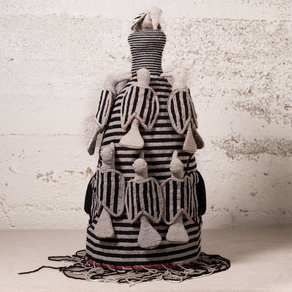 A handmade African-style textile doll or decorative sculpture made from black and white striped woven fabric, with small beige fabric figures attached to the sides and a fringed base, set against a textured off-white wall.