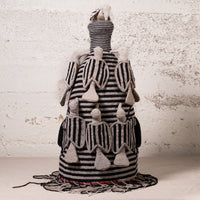A handmade African-style textile doll or decorative sculpture made from black and white striped woven fabric, with small beige fabric figures attached to the sides and a fringed base, set against a textured off-white wall.