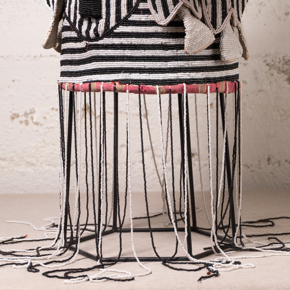 A close-up of a woven textile or rug with horizontal black and white stripes, draped over a black metal hexagonal frame with long hanging black and white cords. A pink patterned band is visible around the upper section, and the background is a textured off-white wall.