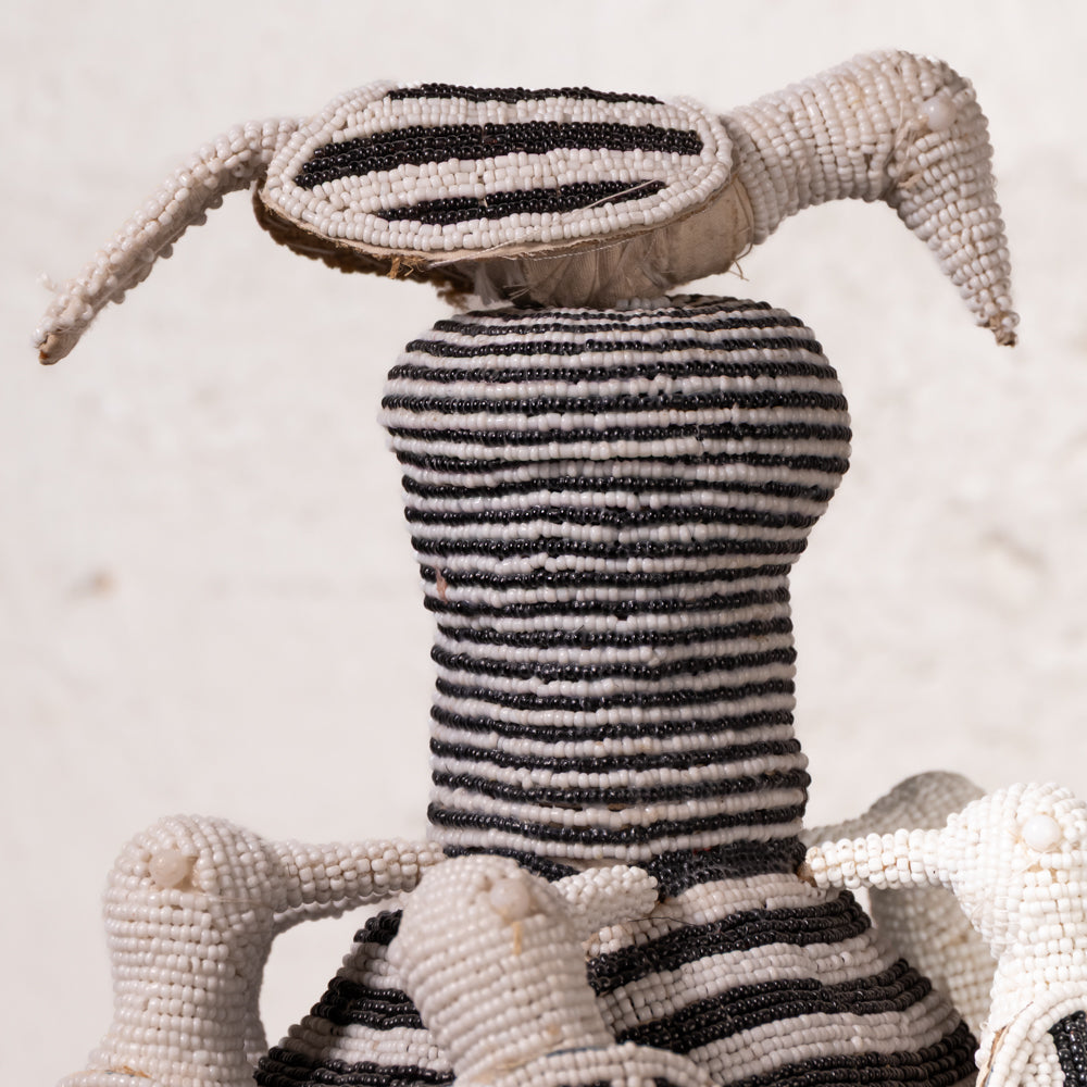A handmade African beaded doll with a striped black and white body, a large striped head, and long pointed ears, shown against a neutral light background.