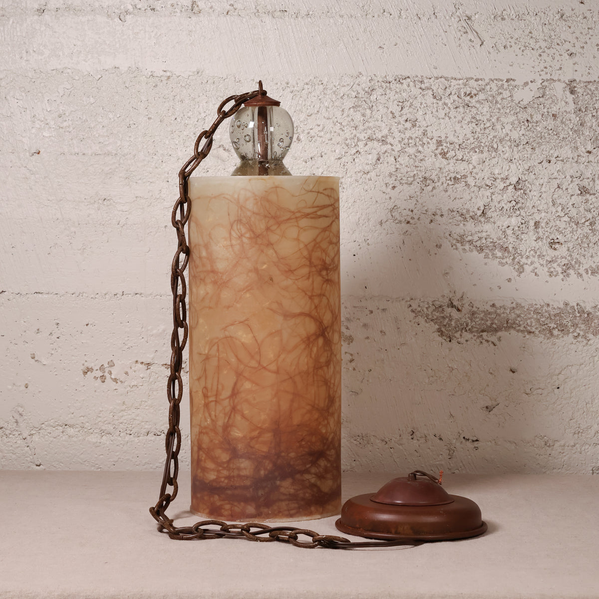 A vintage-style table lamp with a cylindrical amber-brown marbled stone or resin column, a dark metal chain, a clear glass globe shade, and a matching round base, set against a distressed off-white wall.
