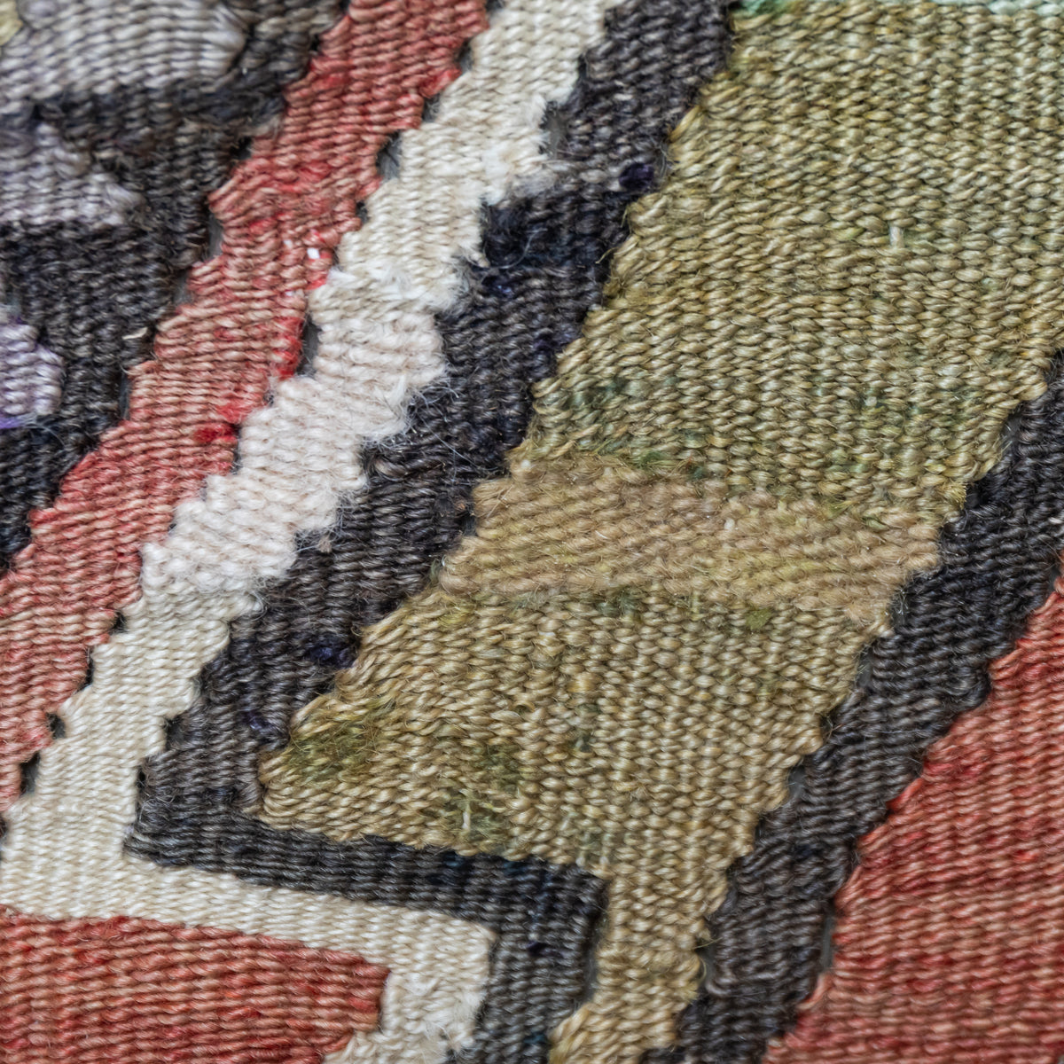 Close-up of a woven textile pattern with diagonal bands in olive green, cream, rust red, charcoal gray, and black.