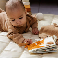 Baby lying on a white blanket with toys around, holding a wooden rattle.