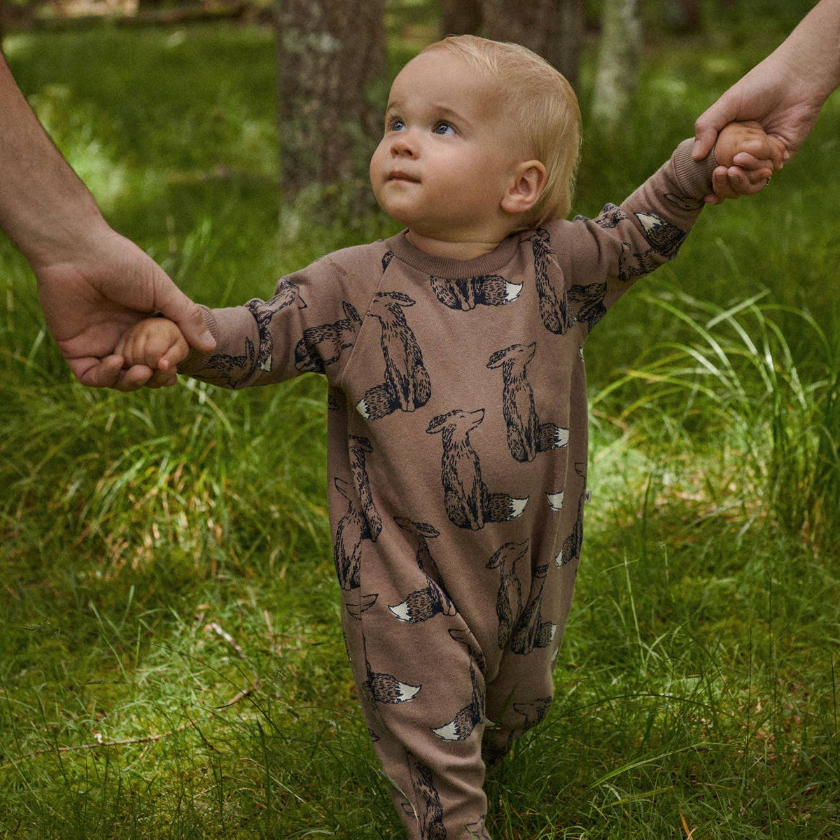 Child wearing a brown onesie with animal patterns, standing in a grassy area.
