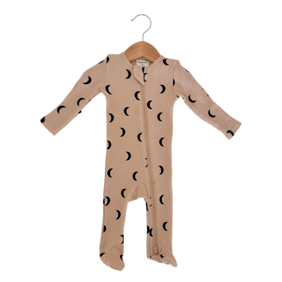 A baby long-sleeve footed sleeper in beige with a black moon phase pattern, hanging on a wooden hanger against a white background.