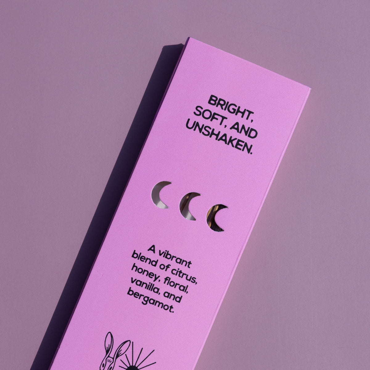 A tall, narrow lavender-pink product box for Brass Earth, featuring black text and illustrations including a rabbit and moon phases, photographed on a matching lavender background.
