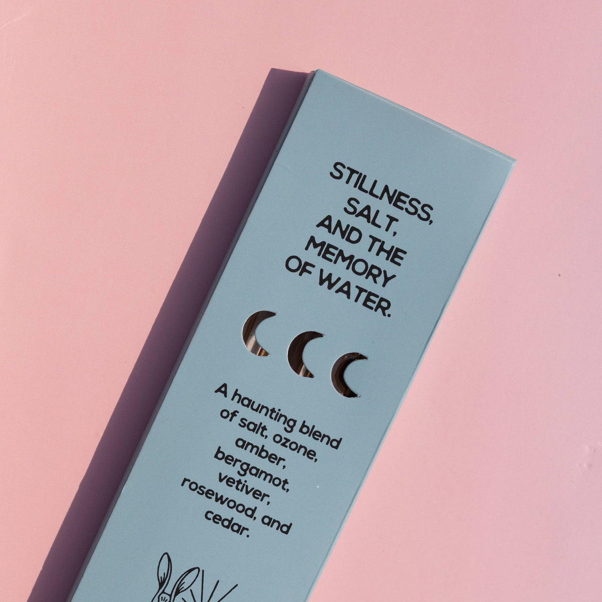 A light blue rectangular product box for Grass Earth essential oils, photographed diagonally on a soft pink background. The box features black text describing a scent profile ('Stillness, Salt, and the Memory of Water') and a simple rabbit illustration.