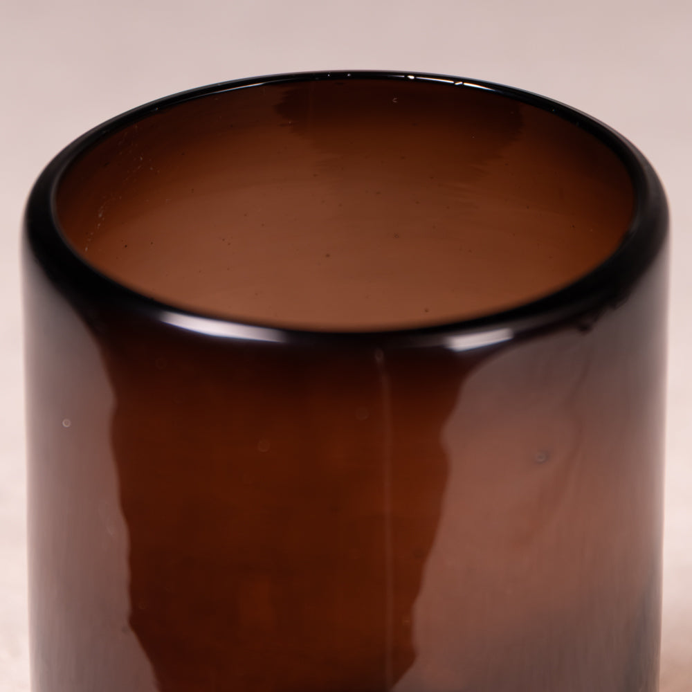 A close-up of a glossy amber brown glass container or vase against a light beige background.