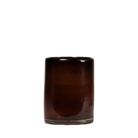 A small cylindrical dark amber glass candle holder or tumbler with a smooth, glossy finish, standing on a light beige surface against a textured, light off-white wall.