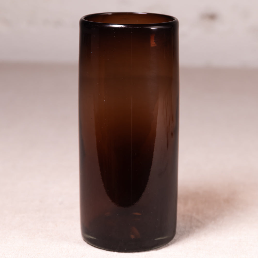 A tall, cylindrical amber brown glass vase with a smooth gradient finish, standing on a neutral light beige surface against a soft, out-of-focus light gray background.