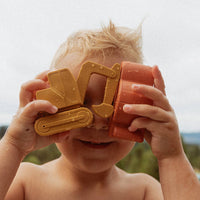 A young child outdoors holding a pair of toy construction binoculars over their eyes. The toy binoculars are made of two colors: one is a warm mustard yellow, and the other is a terracotta orange.