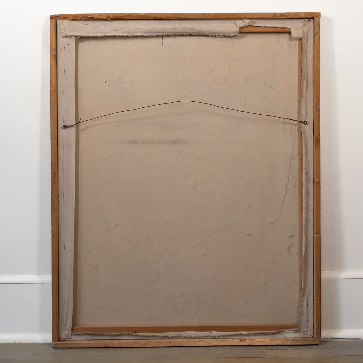 The back view of a large wooden framed canvas or painting, showing the canvas, hanging wire, and wooden stretcher bars, leaning against a white wall.