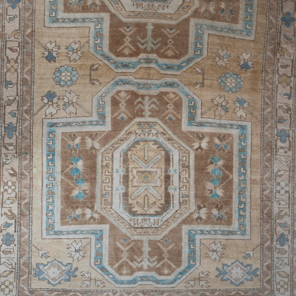 A close-up of a traditional patterned rug with a central medallion, featuring a beige and brown base with light blue and cream accents.