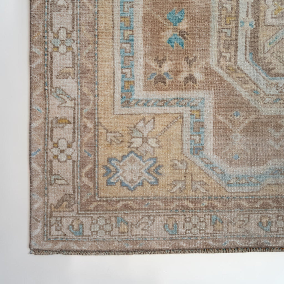 A close-up of a traditional patterned rug corner showing beige, tan, cream, and light blue motifs with a woven texture.