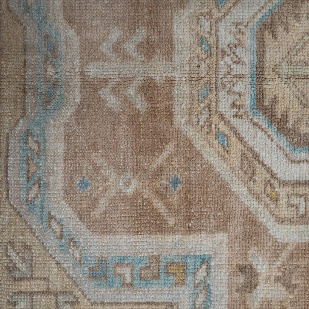 Close-up of a woven rug showing a beige and tan field with geometric motifs in light blue, cream, and small yellow accents.