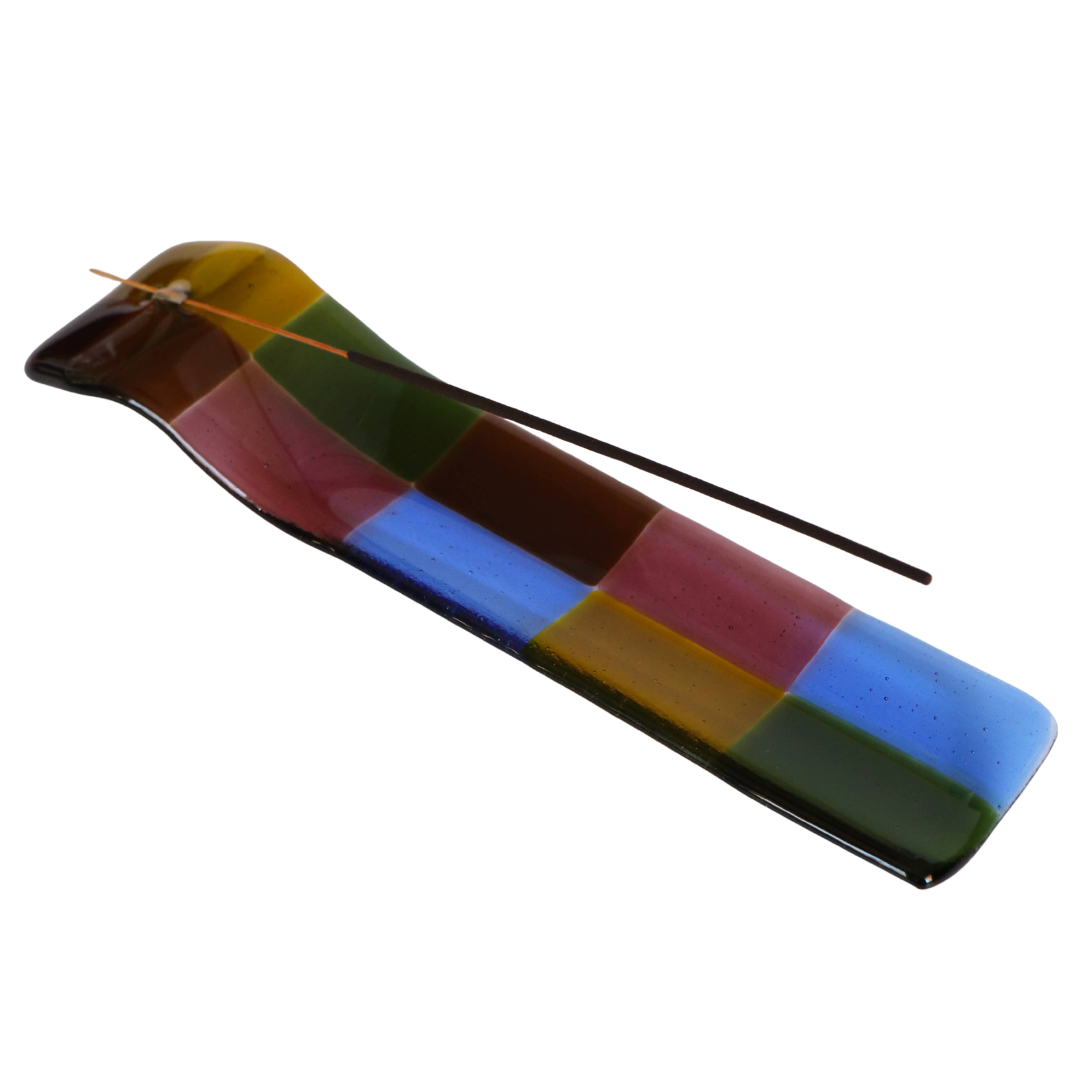 A glossy rectangular glass incense holder with a patchwork design of multiple colors including blue, green, brown, yellow, and red, shown with a single incense stick on a plain white background.