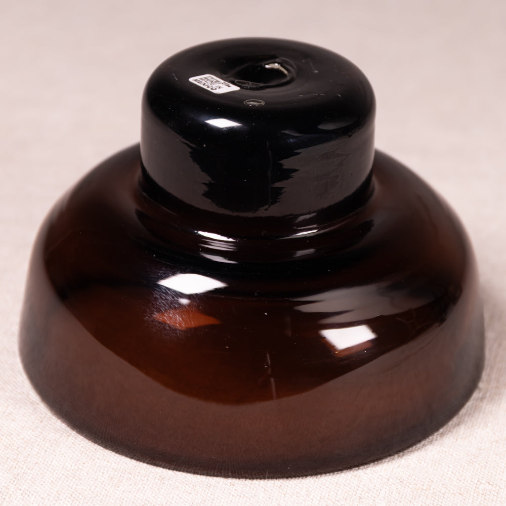 A glossy dark brown glass vessel or container with a wide rounded base and a smaller cylindrical top, photographed on a light beige fabric background.