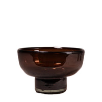 A small amber-brown translucent glass bowl with a pedestal base, photographed against a softly blurred light neutral background.