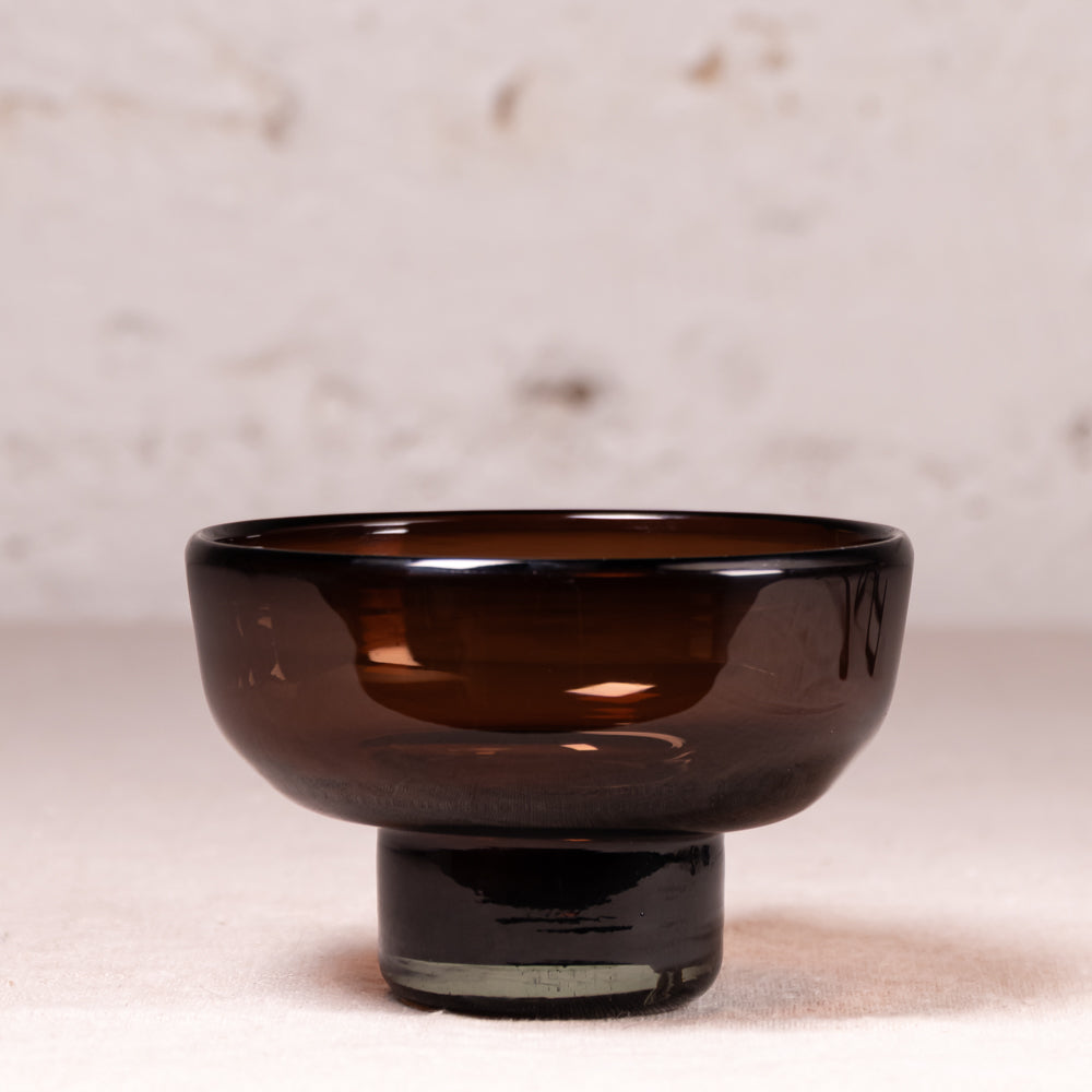 A dark amber brown glass bowl with a matching cylindrical pedestal base, set against a light beige textured background.