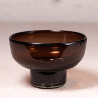 A dark amber brown translucent glass footed bowl with a glossy finish, set against a neutral beige background.