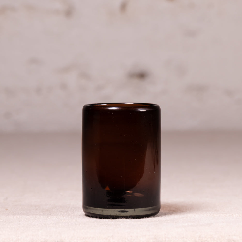A small cylindrical amber glass tumbler or candle holder with a thick base, standing on a light beige textured surface against a soft, out-of-focus neutral background.