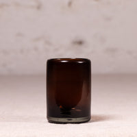A small cylindrical amber glass tumbler or candle holder with a thick base, standing on a light beige textured surface against a soft, out-of-focus neutral background.
