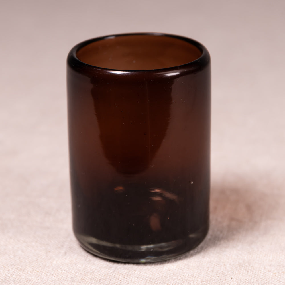 A small cylindrical amber brown glass tumbler or candle holder, photographed on a neutral beige fabric background.