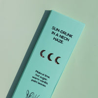 A mint green rectangular product box for Brass Earth, photographed at an angle on a matching mint background. The box features black text reading 'SUN-DRUNK IN A NEON HAZE.