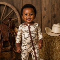 A smiling baby stands barefoot in a rustic western-themed setting, wearing a cream zip-front onesie with brown horse silhouettes and brown trim. The background includes a wooden wall, a wagon wheel, a saddle, a hay bale, a cowboy hat, and a cowhide rug.