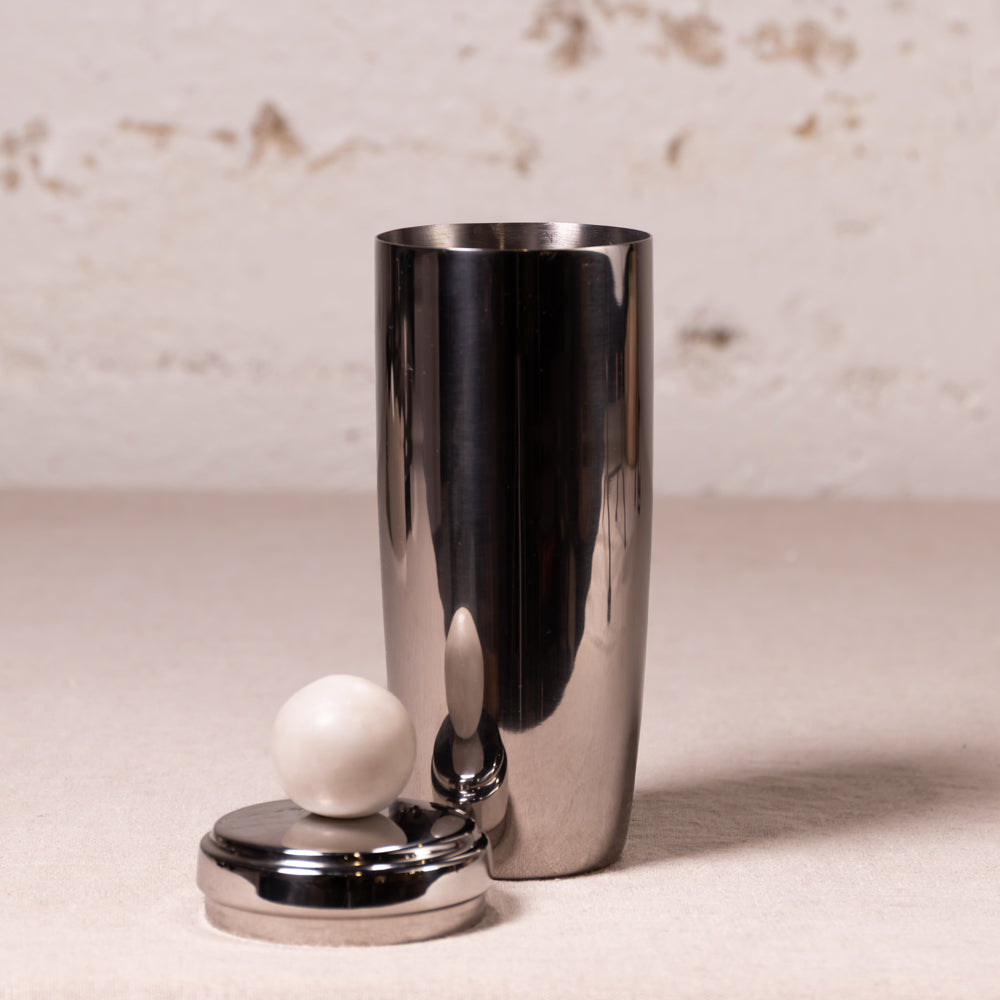 A polished stainless steel cocktail shaker with its metal lid and white marble sphere stopper placed beside it on a light beige surface against a textured off-white wall.
