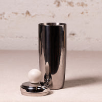 A polished stainless steel cocktail shaker with its metal lid and white marble sphere stopper placed beside it on a light beige surface against a textured off-white wall.