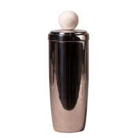A polished stainless steel cocktail shaker with a matching metal lid and a round white wooden stopper on top, set against a neutral, light textured background.