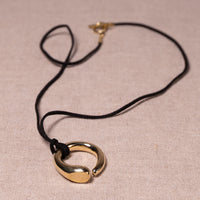 A gold-toned metal pendant necklace with a black cord, displayed on a textured beige fabric background.
