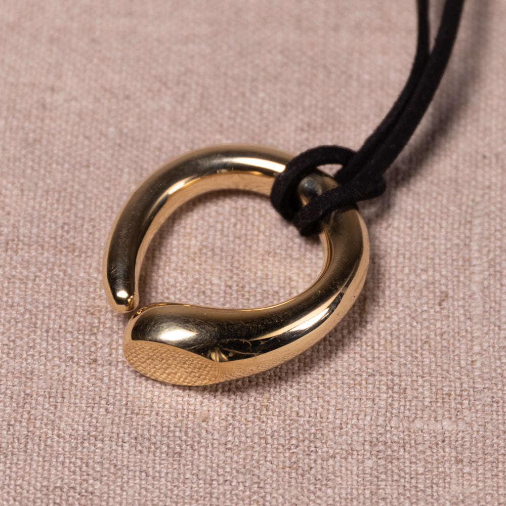 A polished gold-toned metal pendant with a textured finish and a smooth curved end, suspended on a black cord against a beige woven fabric background.