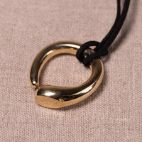 A polished gold-toned metal pendant with a textured finish and a smooth curved end, suspended on a black cord against a beige woven fabric background.